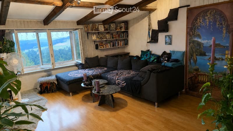 Cozy living room with exposed wooden beams, large windows offering a scenic view, a sectional sofa, coffee table, bookshelf, and decorative items.
