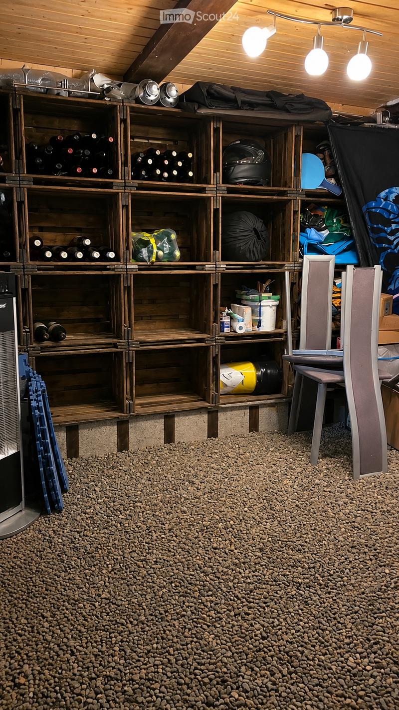 A storage room with wooden shelves filled with wine bottles, helmets, and various storage items. The floor is covered with gravel, and there is a chair near the shelves.