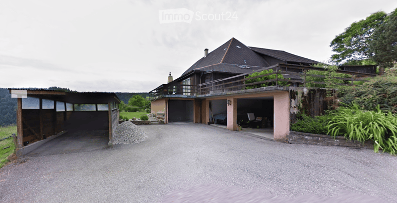 Two-story house with a garage, balcony, and driveway. The house has a dark roof and is surrounded by greenery.