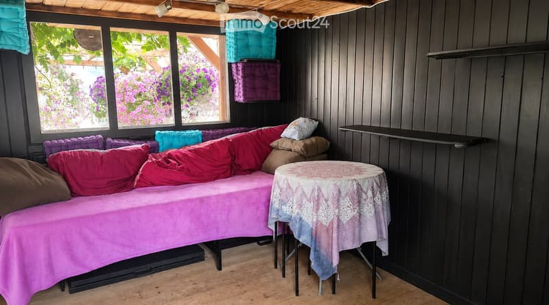 Cozy, wooden-walled room with a large window overlooking a garden, featuring a pink sofa with colorful pillows, a small round table with a decorative tablecloth, and additional cushions on the wall.