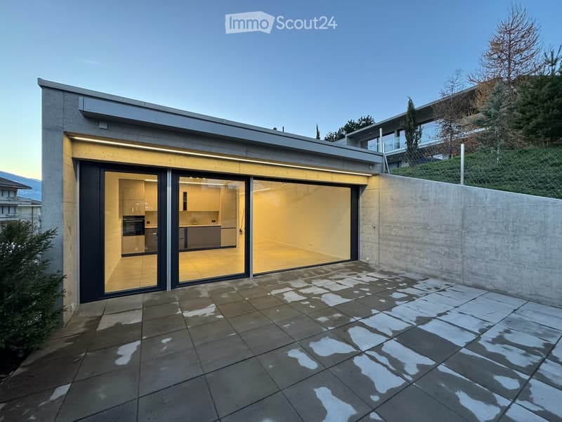 Modern single-story building with large glass sliding doors leading to a tiled patio area. The interior shows a kitchen with cabinets and appliances.