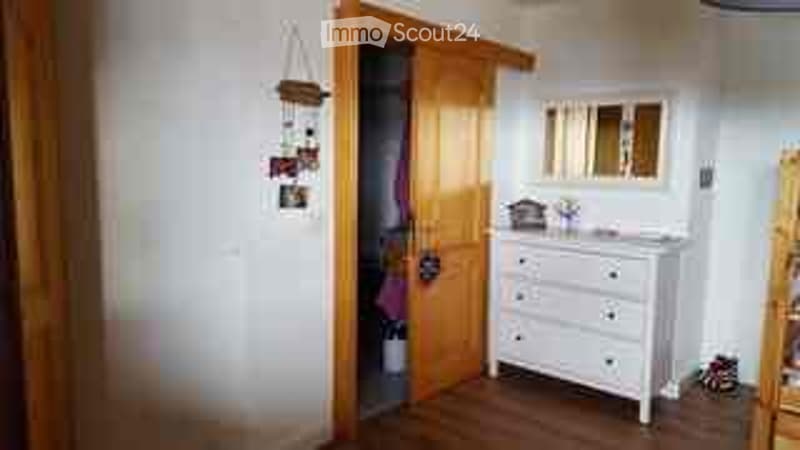 A small, well-lit room with a white dresser, a mirror, and a wooden chair. The room has wooden flooring and white walls.