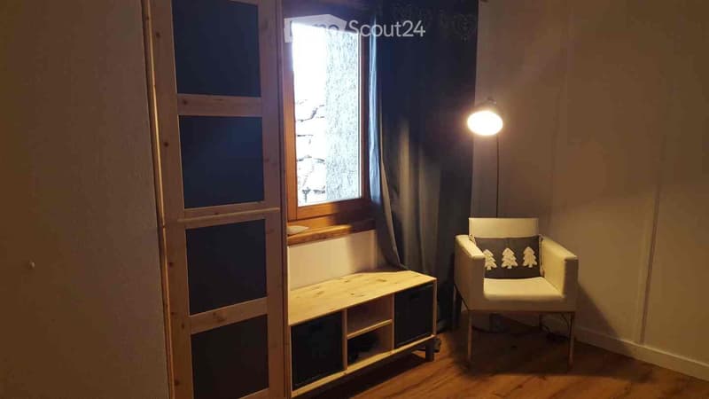 A small, cozy room with wooden flooring, a white armchair with a decorative pillow, a small wooden table, and a window with a curtain. A floor lamp is positioned near the chair.