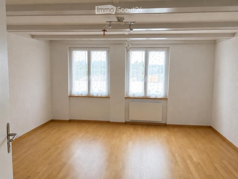 Empty room with wooden flooring, white walls, and large windows with blinds. A radiator is visible under one window, and a door is on the left side.