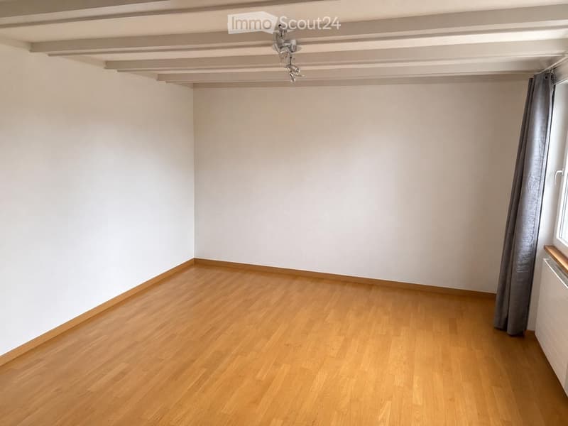 Empty room with light-colored walls and wooden flooring, featuring a window with a curtain and a ceiling light fixture.