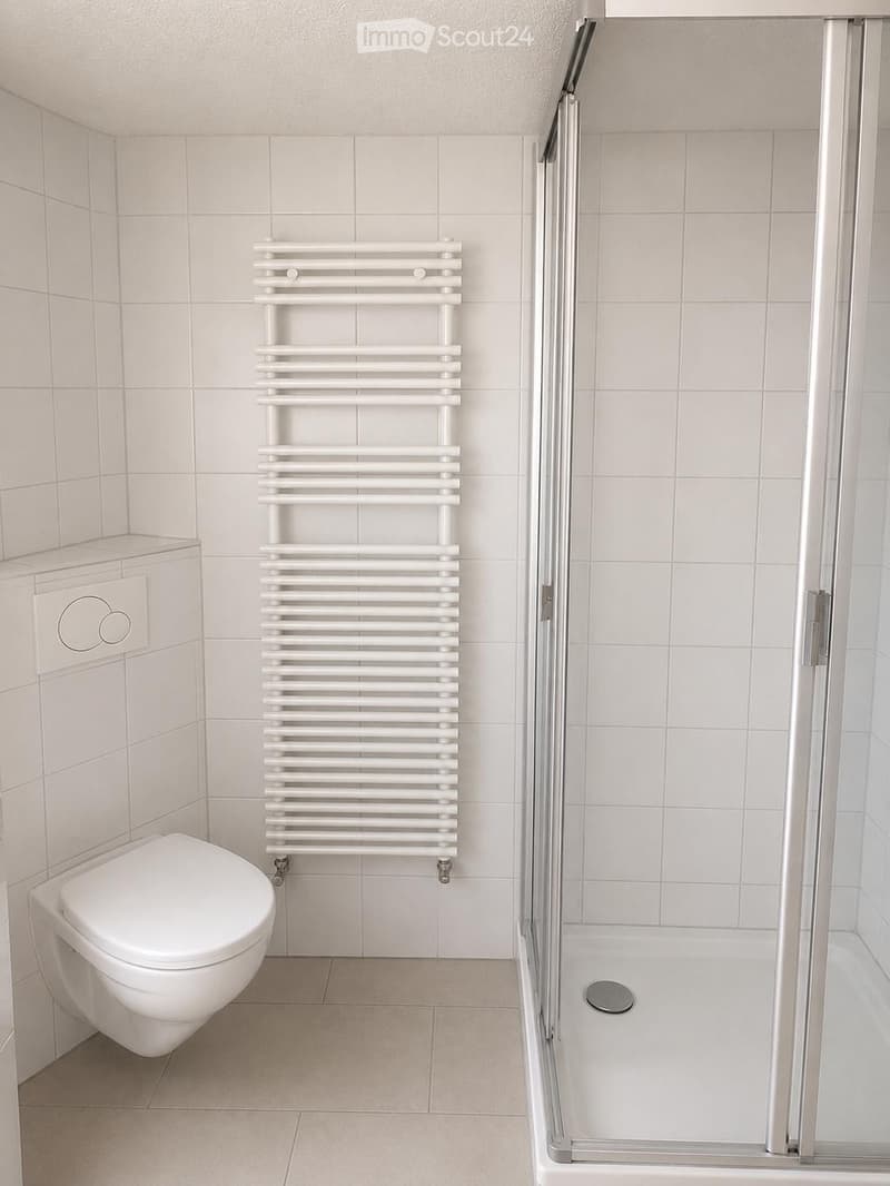 Modern bathroom with white tiled walls and floor, featuring a toilet, a shower enclosure with glass doors, and a white radiator.