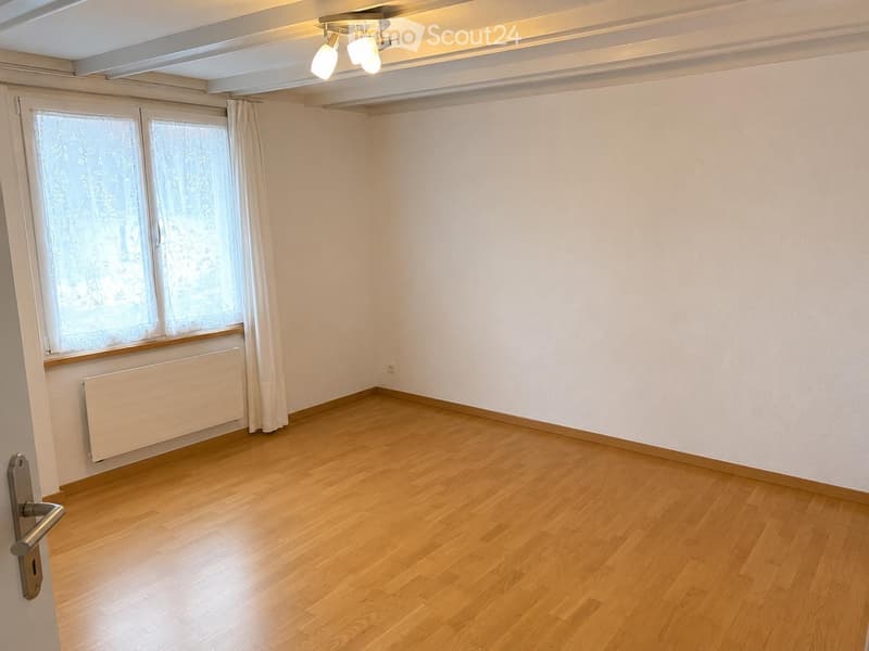 Empty room with wooden flooring, white walls, large window with curtains, radiator, and ceiling light fixture.