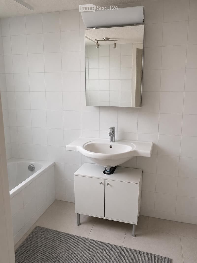 Modern bathroom with white tiled walls, a white sink with a modern faucet, a mirrored cabinet, a bathtub, and a grey bath mat on the floor.