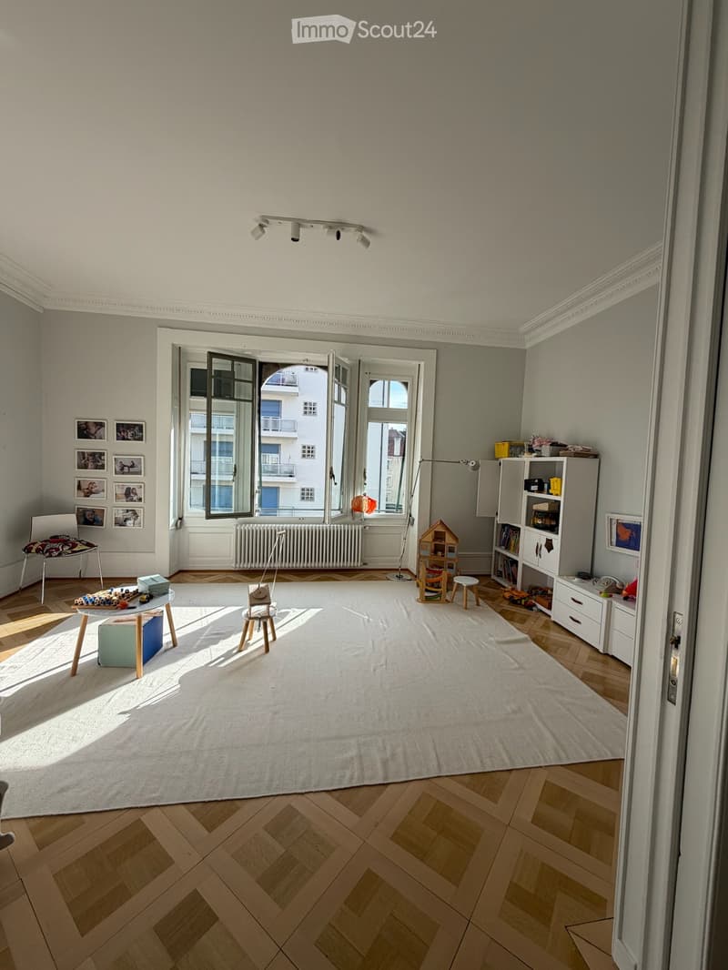 Bright, airy room with wooden herringbone flooring, large white rug, large windows with open shutters, and various children's furniture and toys.