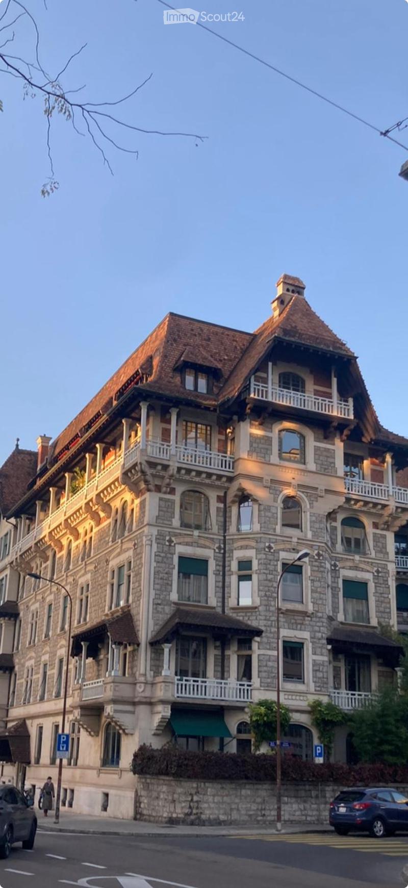 A large, multi-story building with a stone facade, multiple balconies, and a steeply pitched roof. The building features ornate architectural details and is illuminated by warm lighting.