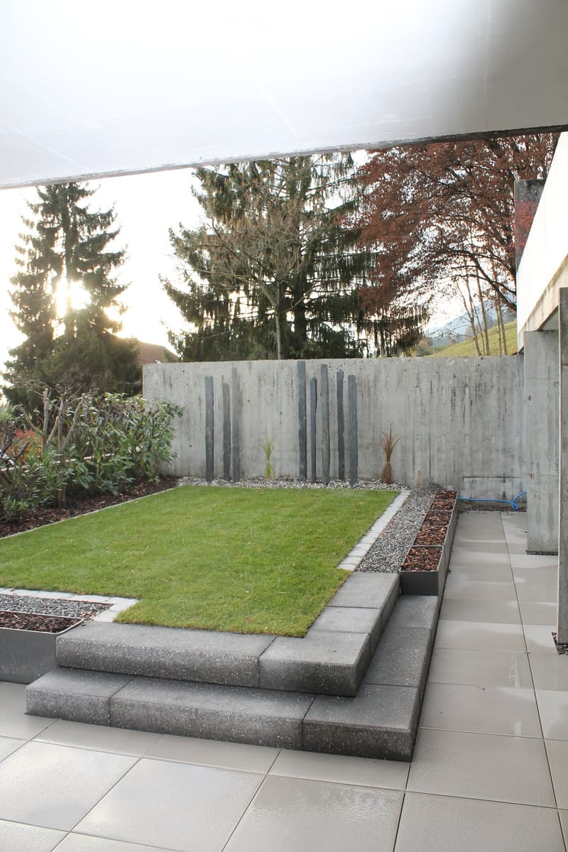 Modern garden with a raised concrete platform, neatly trimmed grass, surrounding trees, and a concrete wall with vertical slats.