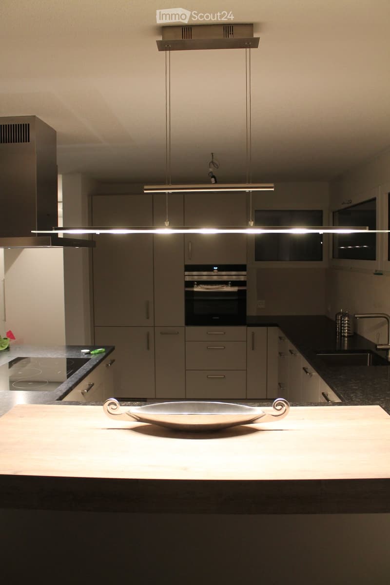 Modern kitchen with grey cabinets, black countertops, and a central island with a wooden cutting board and a decorative bowl. Features include an oven, sink, and pendant lighting.