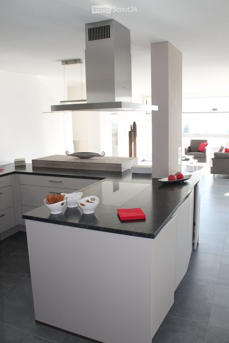 Modern kitchen with white cabinets, dark countertops, and a central island. The island features a built-in induction cooktop and a range hood. The kitchen is open to a living area with a sofa and a coffee table.