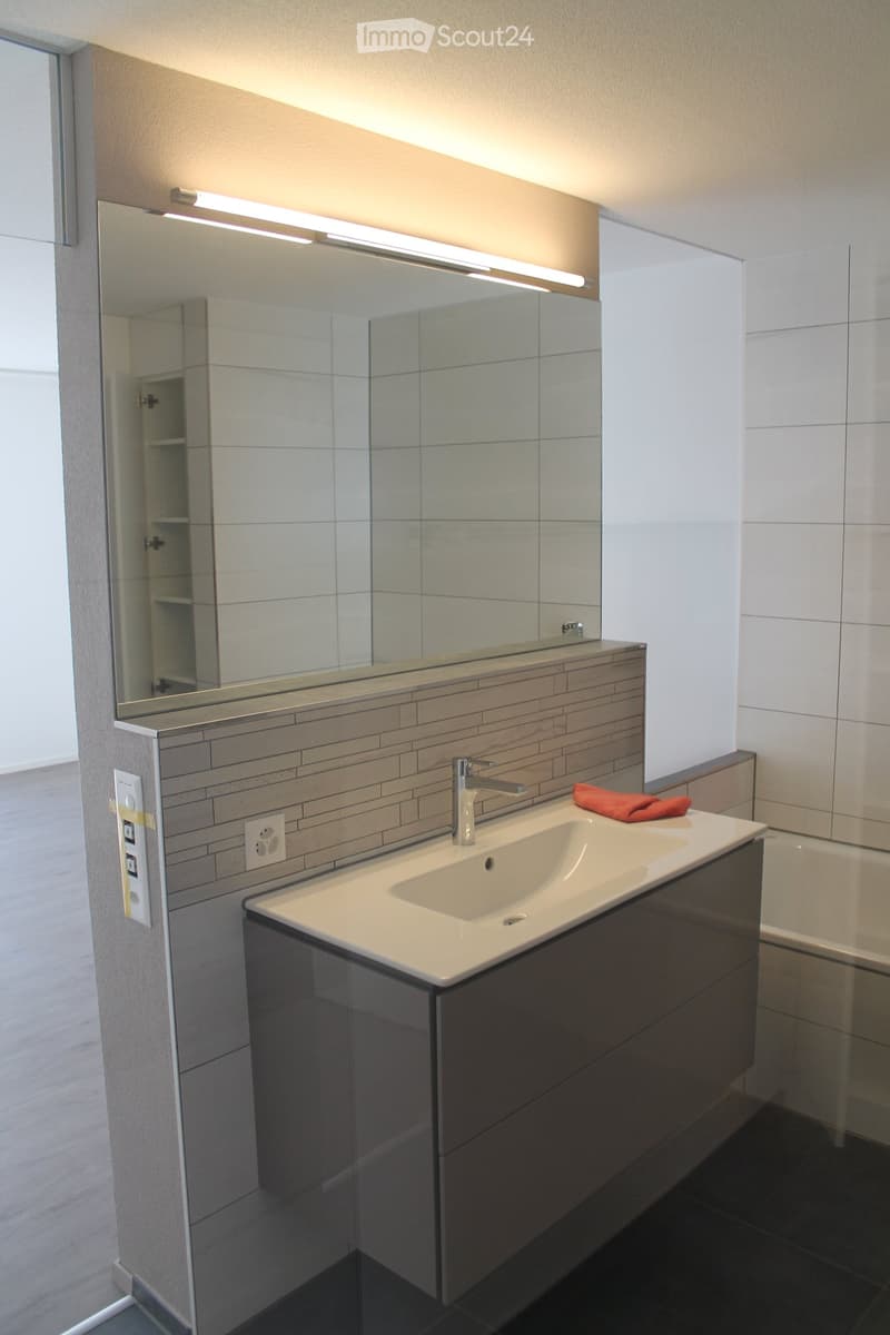 Modern bathroom with a white sink, chrome faucet, large mirror with built-in lighting, dark wooden cabinet, and a red towel on the counter.