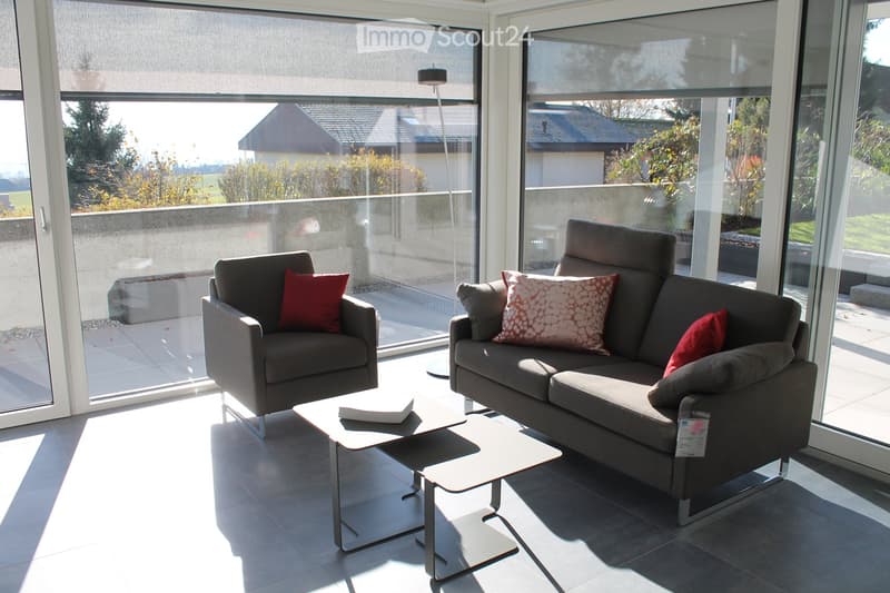 Modern living room with large windows, grey sofa, armchair, coffee table, and floor lamp.