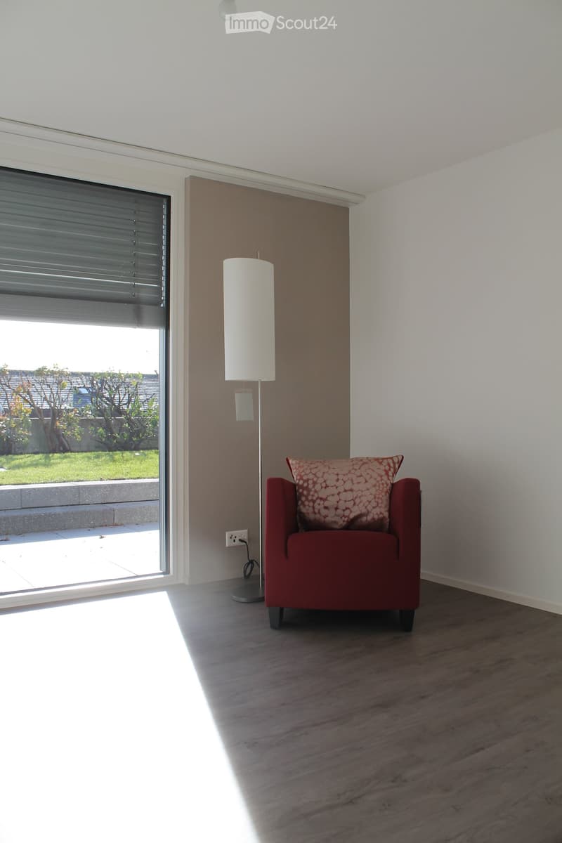 A modern, minimalist room with wooden flooring, a red armchair with a patterned cushion, and a tall floor lamp near a large window with blinds.