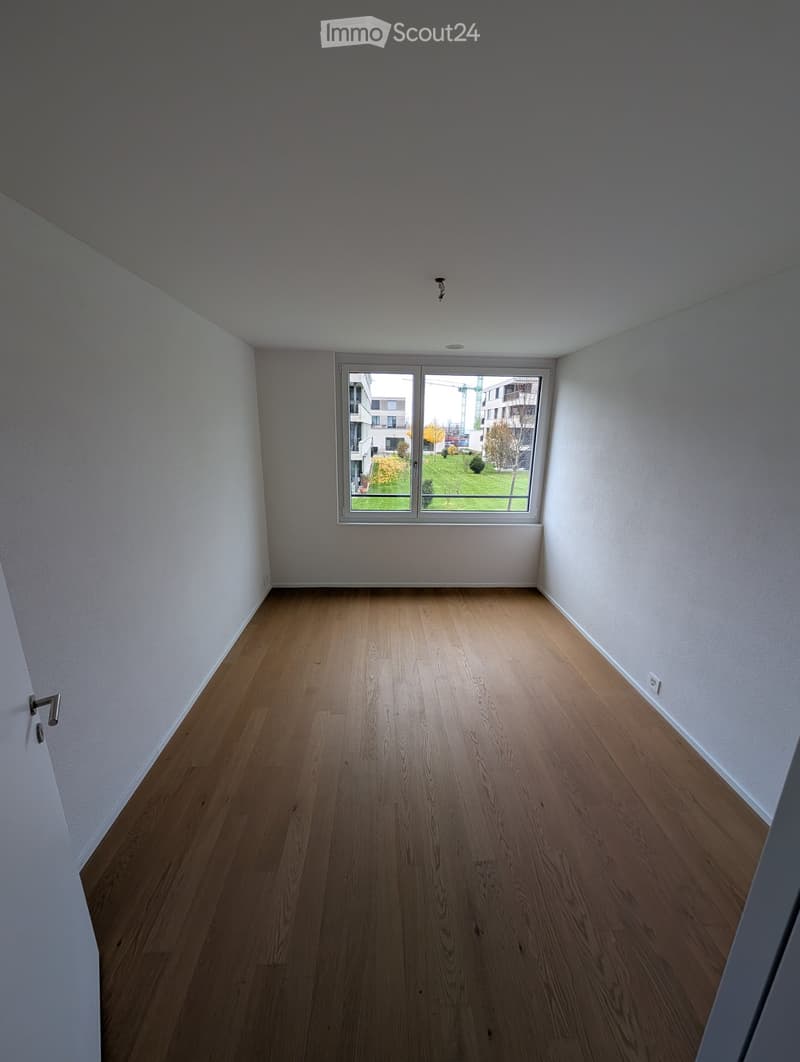 Empty room with wooden flooring, white walls, and a large window overlooking a green area.