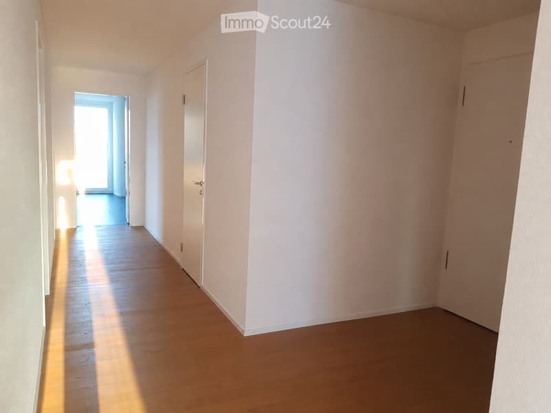Empty hallway with light wooden flooring, white walls, and a closed door on the right side. Natural light streams in from an open doorway at the end of the corridor.