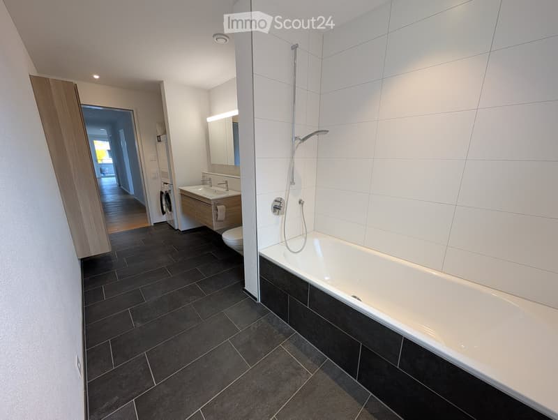 Modern bathroom with dark tiled floor, white bathtub, sink, mirror, and showerhead. A washing machine is visible in the adjacent room.