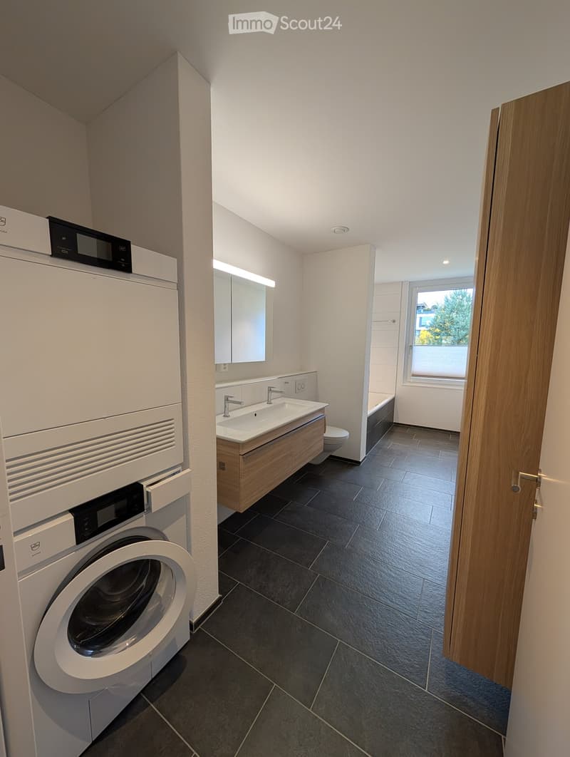 Modern bathroom with dark tiled floor, white walls, a washing machine, a sink with a wooden cabinet, a mirror, a toilet, and a bathtub. A window provides natural light.