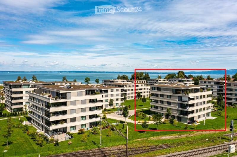 Modern multi-story residential buildings with flat roofs and balconies, surrounded by green lawns and trees, overlooking a body of water.