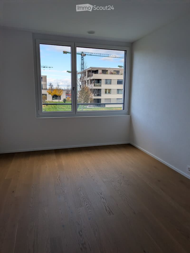 Empty room with wooden flooring and large windows offering a view of a construction site and nearby buildings.