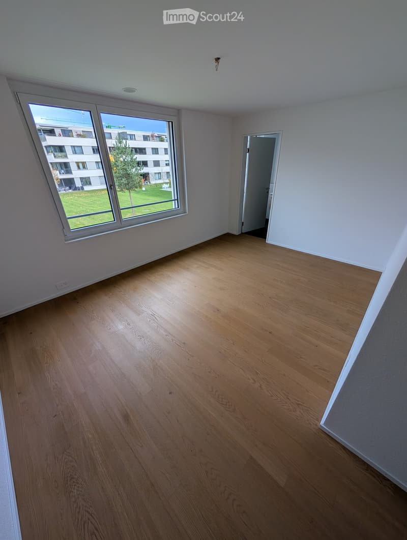 Empty room with wooden flooring, white walls, large window with view of a green area, and a door leading to another space.