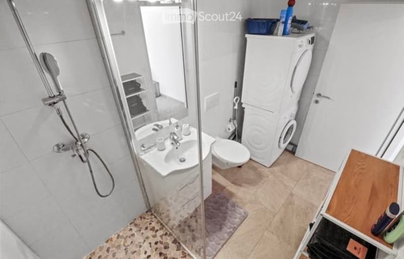 Modern bathroom with a white sink, toilet, glass-enclosed shower, washing machine, and a small storage cabinet. The floor is tiled, and the walls are white.