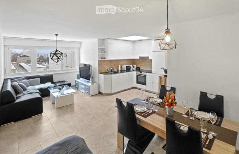 Open-plan living and dining area with modern furniture, including a black sofa, glass coffee table, flat-screen TV, white kitchen units, and a wooden dining table set for six.