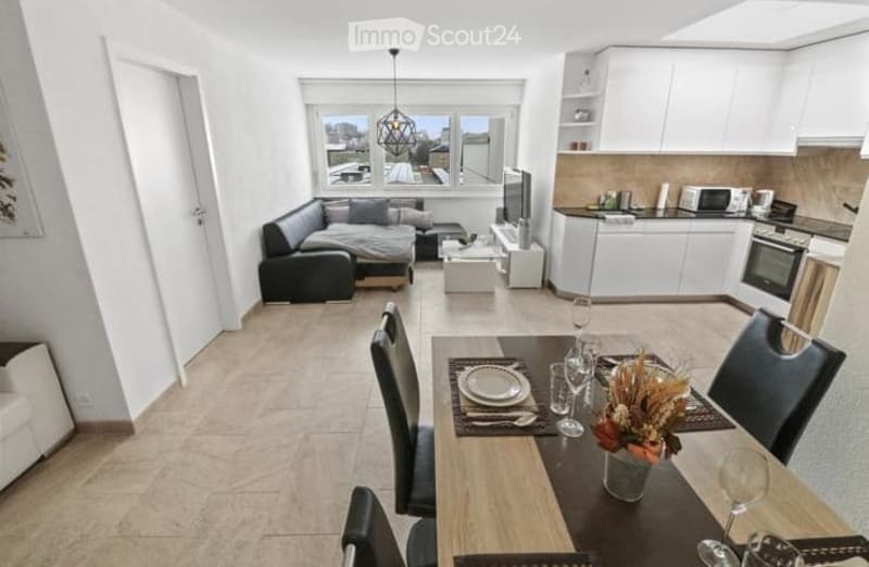 Modern, open-plan living and dining area with a sofa, coffee table, TV stand, dining table with chairs, and a fully equipped kitchen featuring white cabinets, a microwave, and an oven. Large windows offer a view of the city.