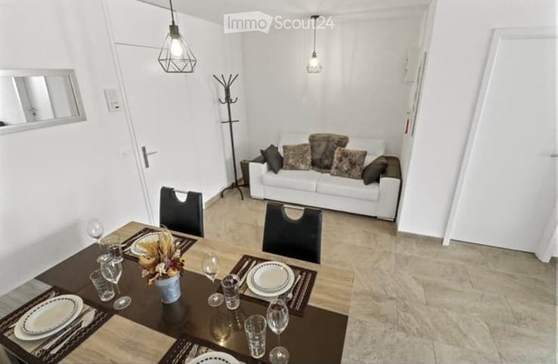 A modern, open-plan living and dining area with a white sofa, dining table with chairs, and decorative items on the table. The space features white walls, pendant lights, and a coat rack near the door.