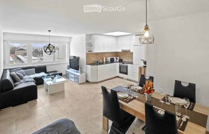 Modern, open-plan living and dining area with a black sofa, glass coffee table, flat-screen TV, sleek white kitchen with cabinets and appliances, and a wooden dining table set with plates, glasses, and a flower vase.