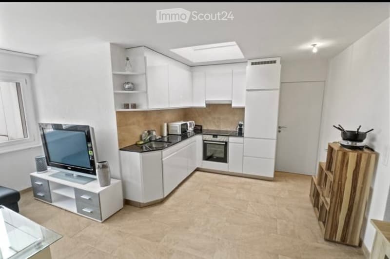 Modern, compact kitchen and living area with white cabinets, a microwave, oven, sink, and a television on a stand. The space includes a small wooden stool and a glass table.