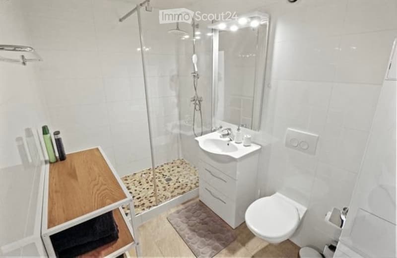 Modern bathroom with white tiles, a glass-enclosed shower, a white sink with a mirror, a toilet, and a small wooden shelf holding towels and toiletries.