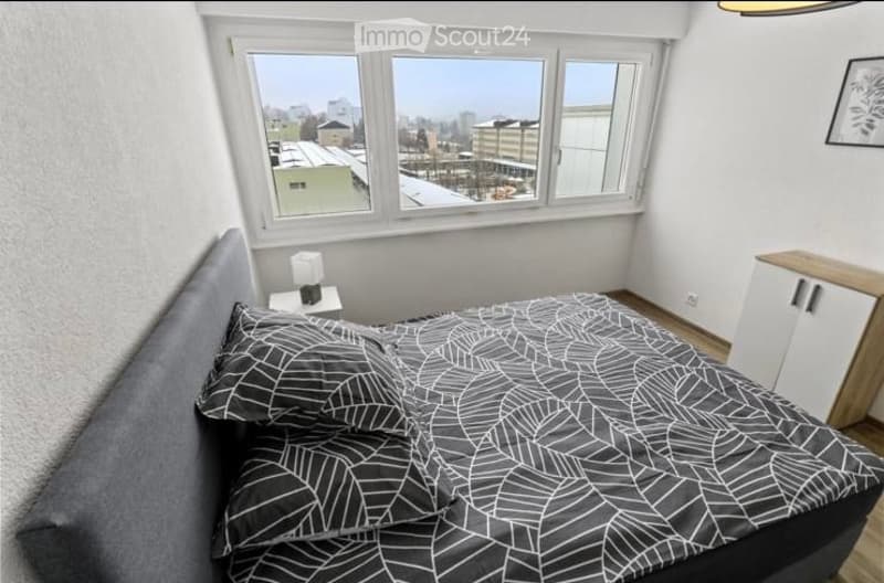 Modern bedroom with large windows offering a city view, featuring a grey upholstered bed with a black and white leaf-patterned duvet, a nightstand with a lamp, and a white cabinet with wooden trim.