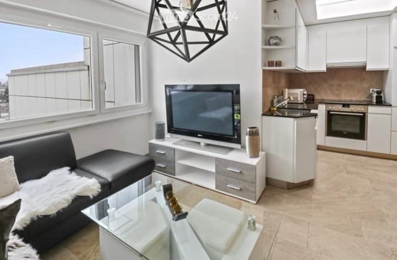 Modern, open-plan living and dining area with a black leather sofa, glass coffee table, flat-screen TV on a white stand, and a compact white kitchen with cabinets, sink, and oven. Large windows offer a city view.