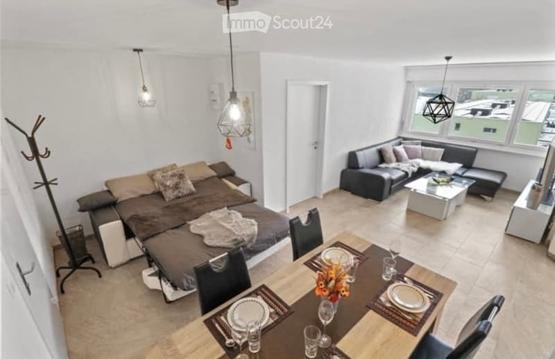 A modern, open-concept living and dining area with a sofa, coffee table, dining table, chairs, and a bed. The space features large windows, pendant lighting, and a neutral color palette.