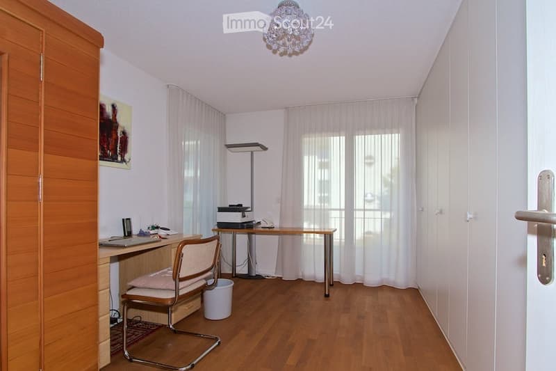 A furnished room with a wooden floor, a desk with a laptop, a chair, a printer, a lamp, a wardrobe, and a picture frame on the wall.