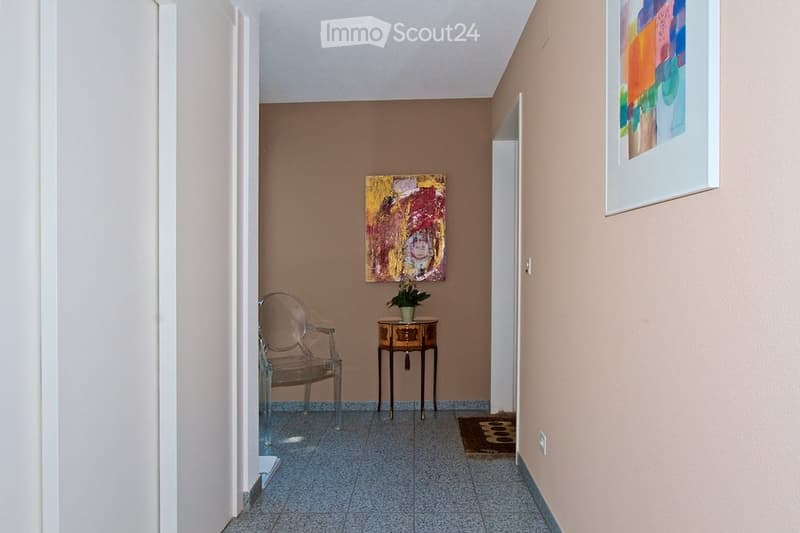 A narrow hallway with beige walls, tiled floor, and two paintings. A small table with a plant and a clear chair are placed on the left side.