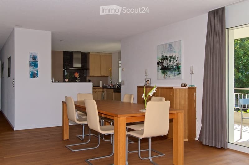 Open-plan dining and kitchen area with wooden dining table, six chairs, modern kitchen cabinets, stove, sink, and decorative items including candles and a picture frame. Large windows and sliding doors lead to a balcony.
