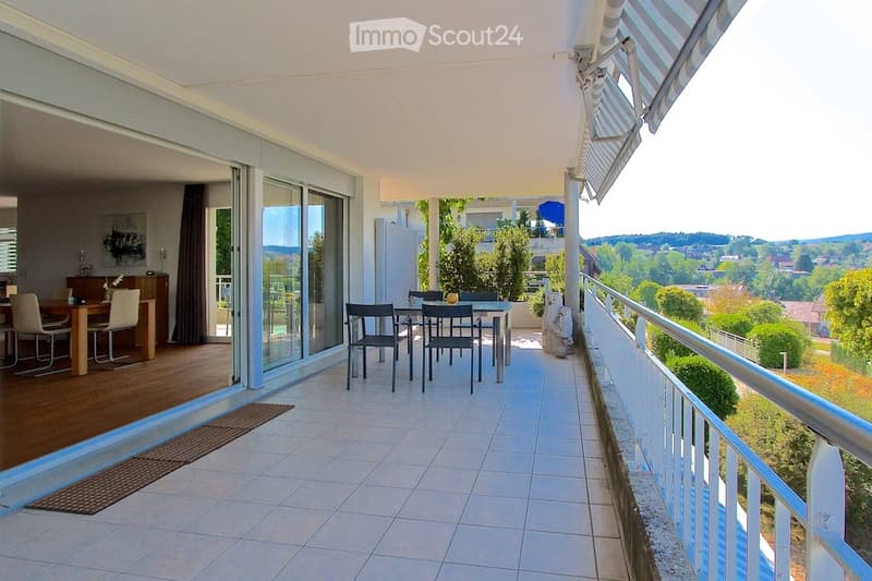 Spacious balcony with tiled floor, glass sliding doors leading to a dining area inside, outdoor dining set, and scenic view of greenery and hills.