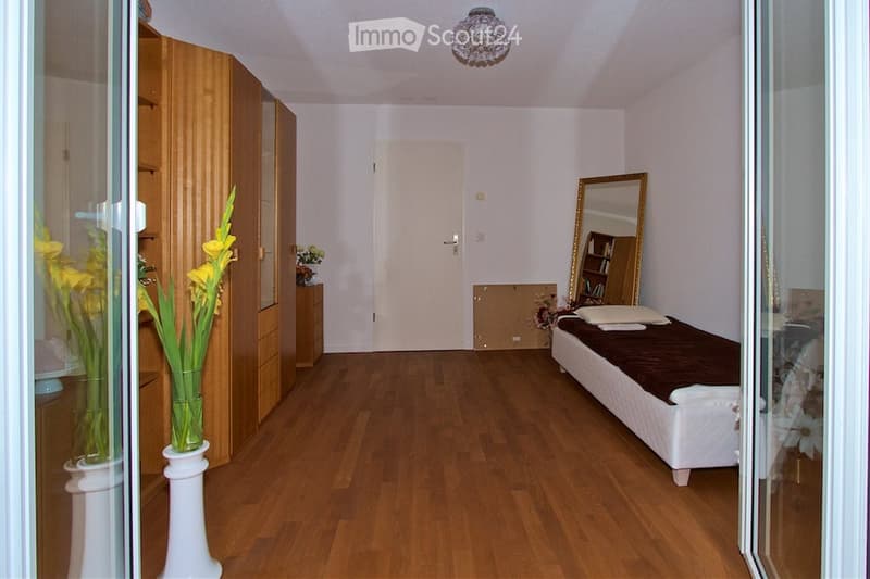 A well-lit bedroom with wooden flooring, a white bed with a brown blanket, a large wardrobe, a mirror, and vases with yellow flowers on a small table.