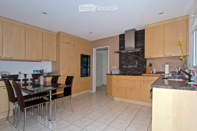 Modern kitchen with light wooden cabinets, granite countertops, stainless steel appliances, and a dining area with a glass table and black chairs.