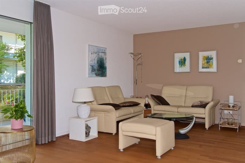 Modern living room with light-colored leather sofas, a glass coffee table, an ottoman, side tables, lamps, and artwork on the walls. Large windows offer a view of the outdoor area.