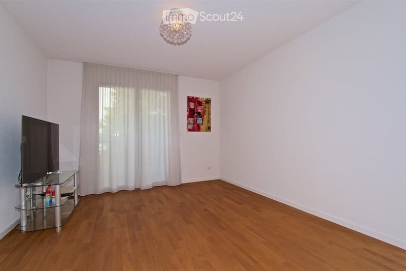 Bright living room with wooden flooring, large window with white curtains, a flat-screen TV on a glass stand, and a decorative picture on the wall.