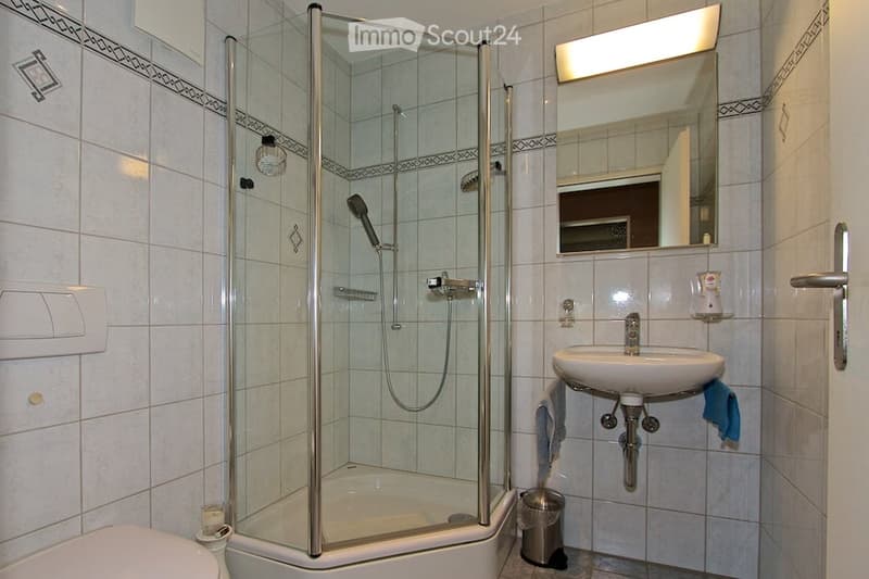 Modern bathroom with a glass-enclosed shower stall, a white sink with a mirror above it, and a toilet. The walls are tiled with a decorative border, and a towel hangs on the side of the sink.