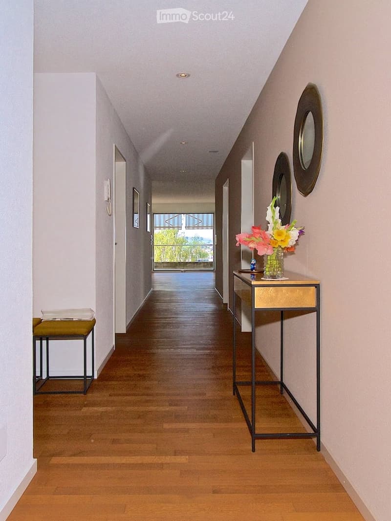 Clean, modern hallway with wooden flooring, white walls, and a small console table with a flower arrangement. There are two decorative mirrors on the wall and a small bench on the left side.