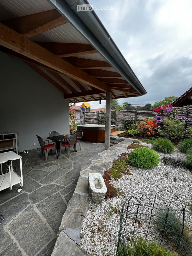 Outdoor patio with stone flooring, covered wooden roof, seating area with table and chairs, hot tub, and garden with colorful flowers and plants.