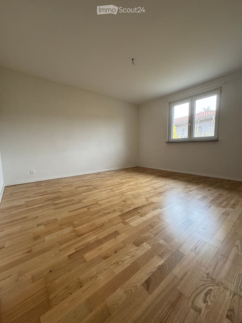 Empty room with light wooden flooring and white walls, featuring a large window with a view of a neighboring building.