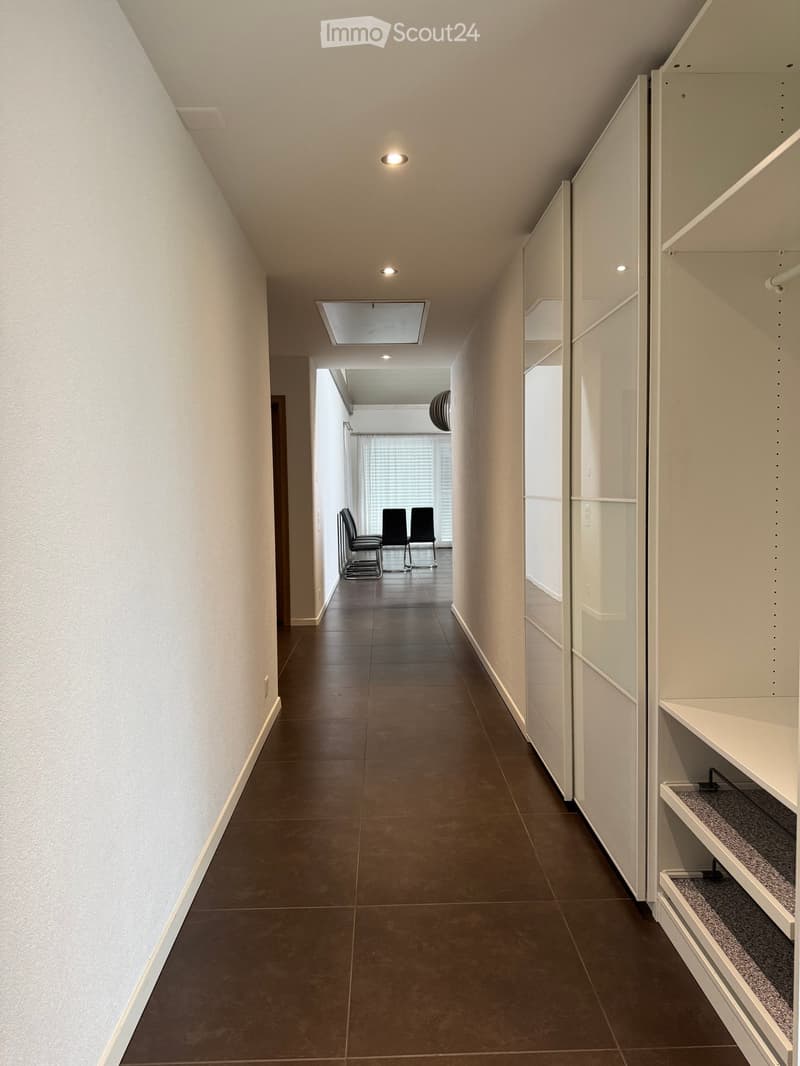 Modern hallway with dark tiled floor, white walls, built-in storage units, and recessed lighting. Three chairs are visible in an adjacent room.